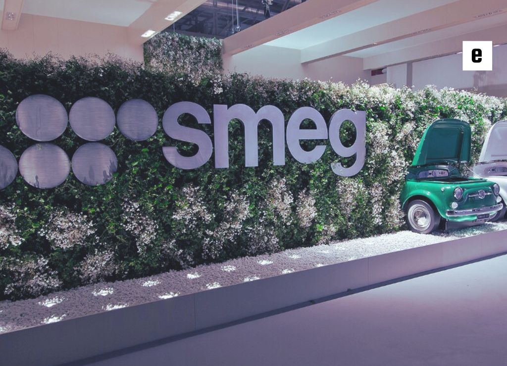 لوگوی برند اسمگ با طراحی سفید روی پس‌زمینه‌ای طبیعی از برگ‌ها و بوته‌های سبز.

